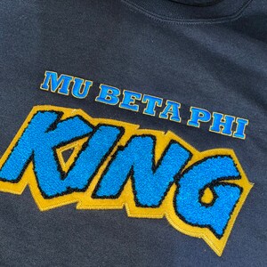 May include: Navy blue sweatshirt with the text "MU BETA PHI KING" in a blue, gold, and black design. The word "KING" is in a large, stylized font. The text "MU BETA PHI" is above "KING".
