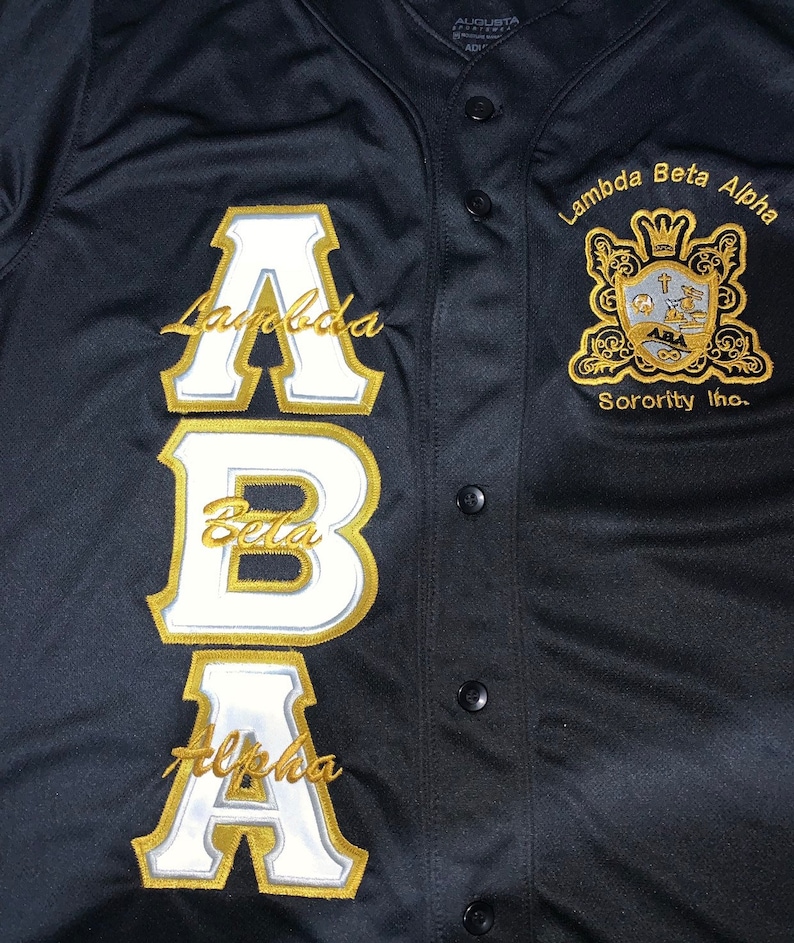 K&ouml;nnte beinhalten: Schwarzes Baseballtrikot mit goldenen und wei&szlig;en gestickten Buchstaben, die "ABA" buchstabieren, und den Worten "Lambda Beta Alpha Sorority Inc." auf der rechten Seite.