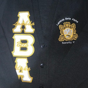 May include: Black cardigan with a gold and white embroidered "ABA" on the left side and a gold embroidered crest with the text "Lambda Beta Alpha Sorority Inc." on the right side.