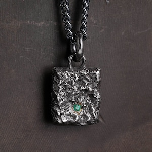 May include: A silver chain necklace with a square pendant. The pendant is textured and has a small green gemstone in the centre.