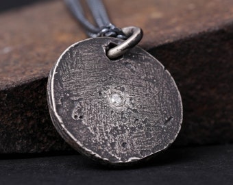 May include: A silver pendant necklace with a round, textured disc featuring a small, clear gemstone in the centre. The necklace is on a black chain.