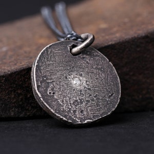 May include: A silver pendant necklace with a round, textured disc featuring a small, clear gemstone in the centre. The necklace is on a black chain.