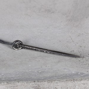 Könnte beinhalten: Eine silberne Halskette mit einem langen, dünnen, spitzen Anhänger. Der Anhänger ist strukturiert und hat einen runden Ring oben.