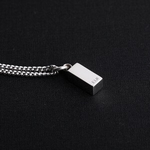 Op de afbeelding: Een zilverkleurige rechthoekige hangerketting met een gourmetteketting. De hanger heeft de letters "EIJI" gegraveerd op de zijkant. De ketting wordt getoond tegen een zwarte achtergrond.