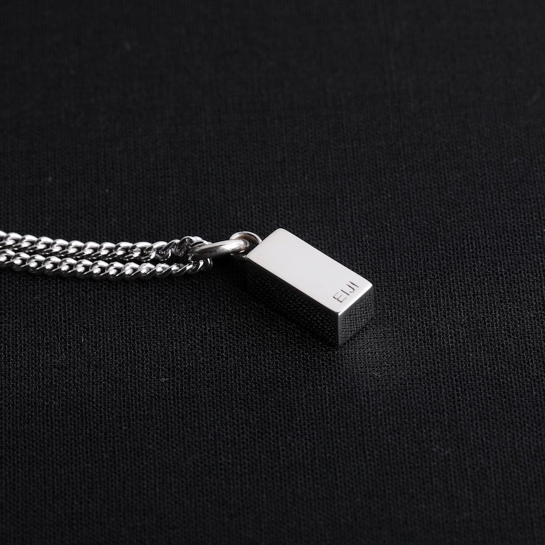 Op de afbeelding: Een zilverkleurige rechthoekige hangerketting met een gourmetteketting. De hanger heeft de letters "EIJI" gegraveerd op de zijkant. De ketting wordt getoond tegen een zwarte achtergrond.