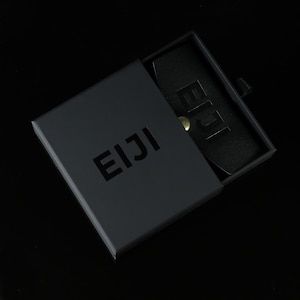 May include: A black box with the word "EIJI" printed on the top. The box is open and a black leather card holder with the word "EIJI" embossed on it is inside.