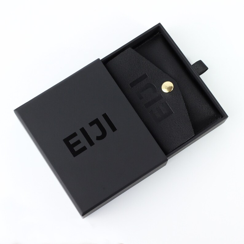 Op de afbeelding: Een zwarte doos met het woord "EIJI" op de voorkant. Binnenin is een zwarte leren portemonnee zichtbaar, vastgezet met een goudkleurige drukknop. Het woord "EIJI" staat ook op de portemonnee.