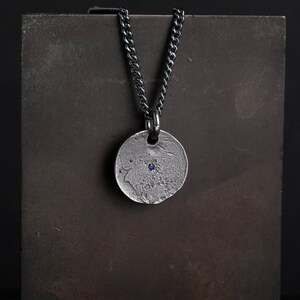 May include: A silver necklace with a round pendant featuring a small blue gemstone. The chain is black and has a curb link design.