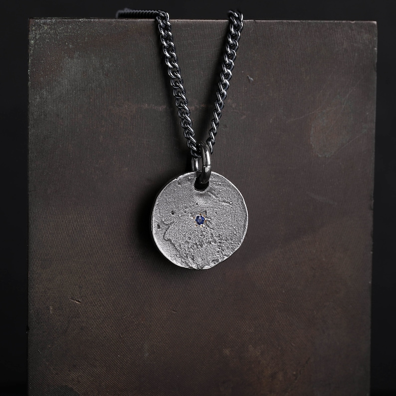 May include: A silver necklace with a round pendant featuring a small blue gemstone. The chain is black and has a curb link design.