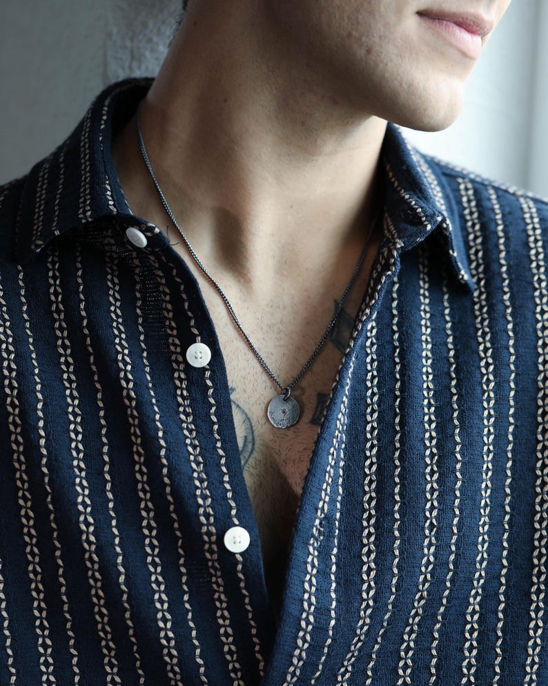 May include: A person wearing a dark blue and white striped shirt with a silver chain necklace and a round pendant.