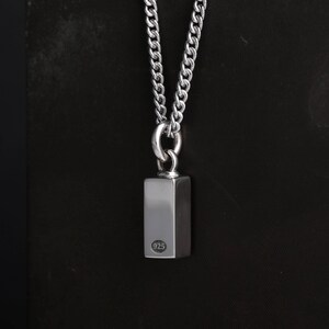 Op de afbeelding: Zilverkleurige ketting met een rechthoekige hanger. De hanger is bevestigd aan een dikke ketting. De hanger heeft een kleine cirkelvormige markering met het nummer "925". Donkere achtergrond.