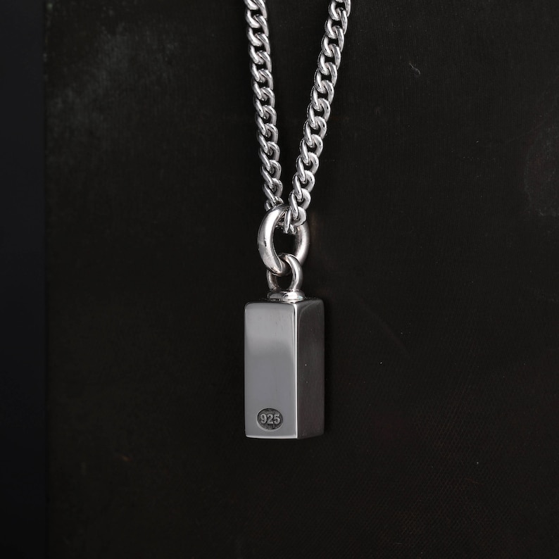 Op de afbeelding: Zilverkleurige ketting met een rechthoekige hanger. De hanger is bevestigd aan een dikke ketting. De hanger heeft een kleine cirkelvormige markering met het nummer "925". Donkere achtergrond.
