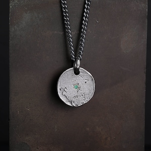 May include: A silver necklace with a round pendant featuring a textured surface and a small green gemstone in the center.