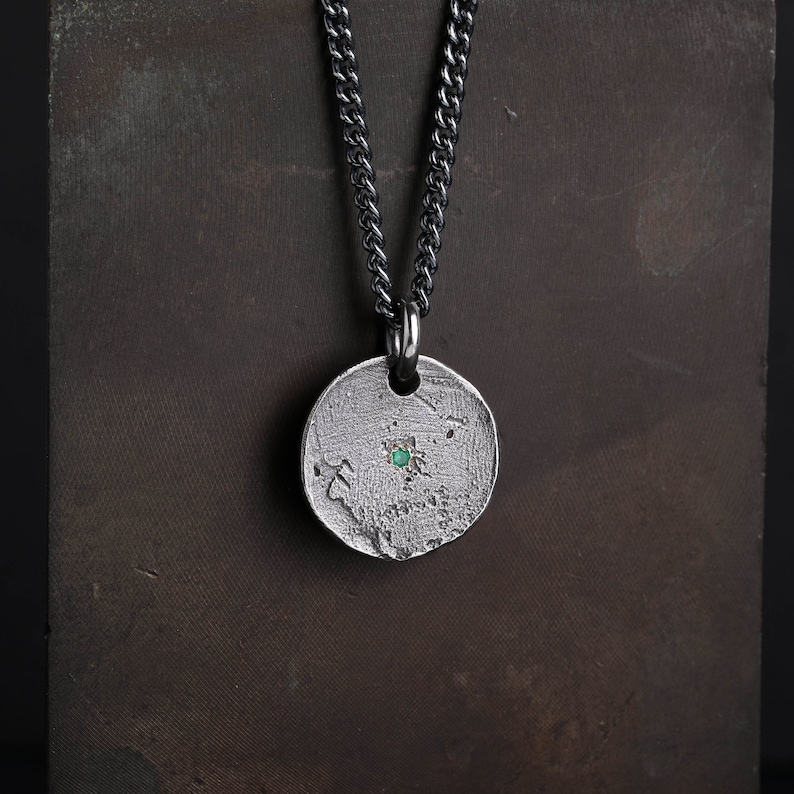 May include: A silver necklace with a round pendant featuring a textured surface and a small green gemstone in the center.