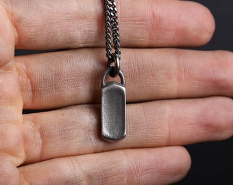 May include: A rectangular, silver-toned pendant necklace with a matching chain. The pendant has a slightly textured surface and a rounded bottom. The chain is a curb style, and the necklace is displayed against a dark background.