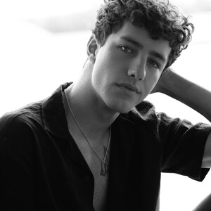 Op de afbeelding: Zwart-wit portret van een persoon met krullend haar, gekleed in een zwart overhemd en een zilveren ketting met een hanger. De persoon kijkt met een ontspannen uitdrukking naar de camera.