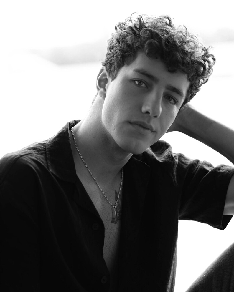 Op de afbeelding: Zwart-wit portret van een persoon met krullend haar, gekleed in een zwart overhemd en een zilveren ketting met een hanger. De persoon kijkt met een ontspannen uitdrukking naar de camera.