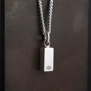 May include: A silver pendant necklace. The pendant is rectangular and polished, hanging from a chain. The pendant has a smooth, reflective surface and a small, circular stamp with the number "925" on the back. The chain has a tightly woven design.