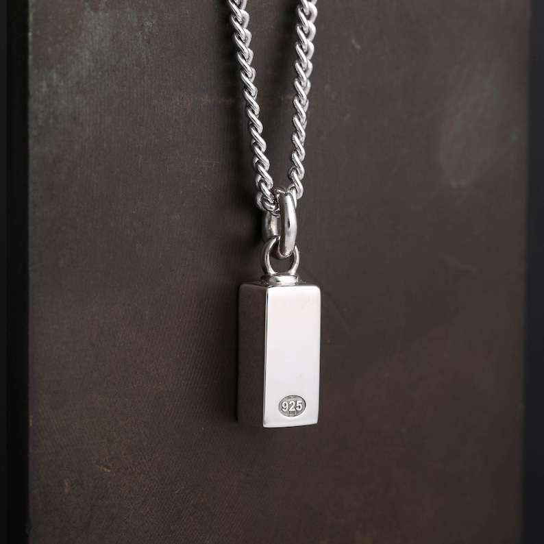 Op de afbeelding: Een zilveren ketting met hanger. De rechthoekige, gepolijste hanger hangt aan een ketting. De hanger heeft een glad, reflecterend oppervlak en een kleine, ronde stempel met het nummer "925" op de achterkant. De ketting heeft een strak geweven ontwerp.