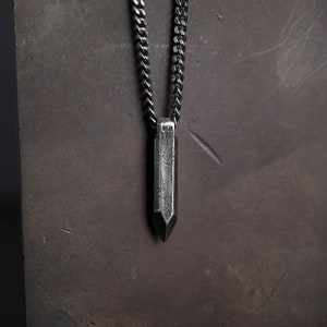 May include: A silver chain necklace with a long, pointed, silver pendant. The pendant has a textured surface and a small inscription.