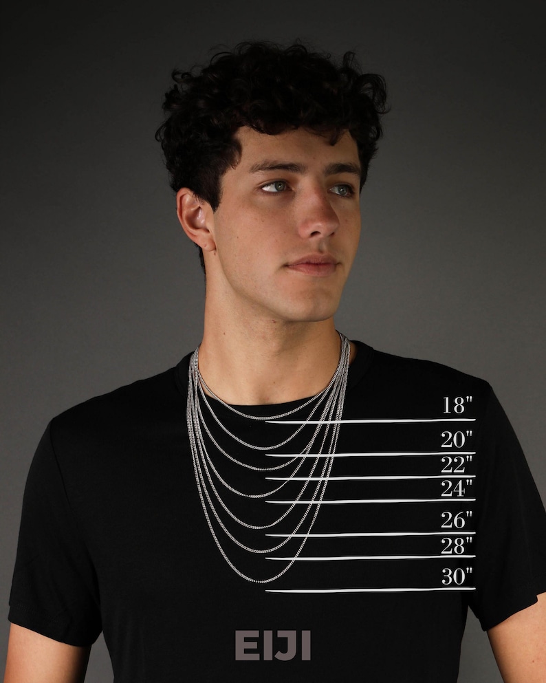 Op de afbeelding: Een persoon draagt een zwart t-shirt met een zilveren ketting. De kettinglengtes worden weergegeven op 45,7 cm, 50,8 cm, 55,9 cm, 61 cm, 66 cm, 71,1 cm en 76,2 cm. De merknaam "EIJI" staat op het shirt.