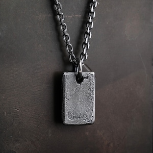 May include: A silver-toned rectangular pendant necklace with a textured surface. The pendant hangs from a matching chain. The necklace is displayed against a dark background.