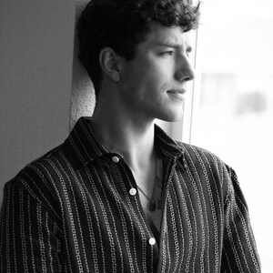 May include: A man with curly hair wearing a striped black and white button-down shirt. He is looking out a window.