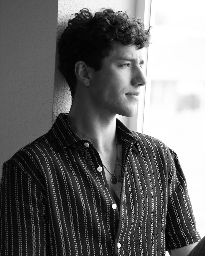 May include: A man with curly hair wearing a striped black and white button-down shirt. He is looking out a window.
