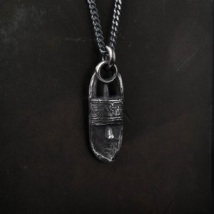May include: A silver-toned pendant necklace with a textured, elongated design. The pendant is suspended from a curb chain. The pendant has a detailed, carved appearance, with a central band and a small, pointed element at the base.