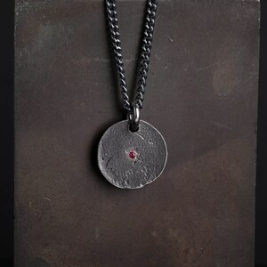 May include: A silver chain necklace with a round pendant featuring a textured surface and a small red gemstone in the center.