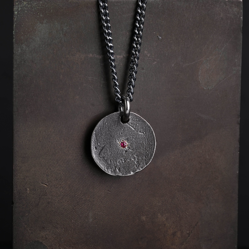 May include: A silver chain necklace with a round pendant featuring a textured surface and a small red gemstone in the center.