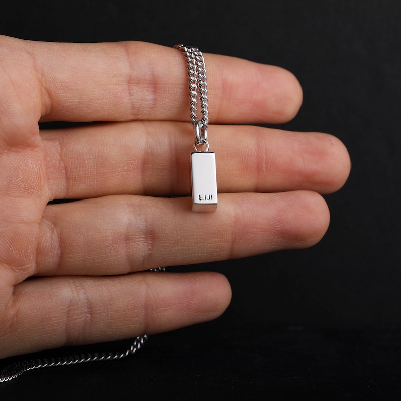 Op de afbeelding: Zilverkleurige ketting met een rechthoekige hanger. Op de hanger staat de tekst "EIJI". De ketting is van het curb-model. De achtergrond is effen zwart.