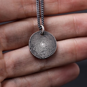 May include: A silver chain necklace with a round pendant featuring a small diamond in the center. The pendant has a textured surface.