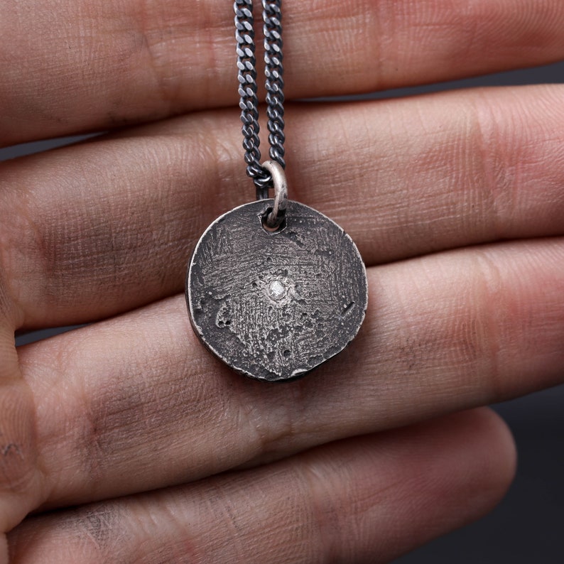 May include: A silver chain necklace with a round pendant featuring a small diamond in the center. The pendant has a textured surface.