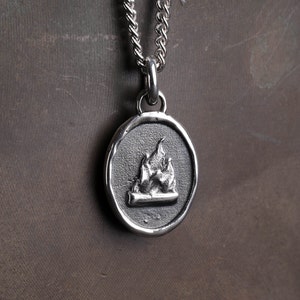 May include: A silver pendant necklace with an oval charm depicting a campfire. The charm shows flames above a log, set against a textured backdrop, and is suspended from a silver chain. This is a piece of jewellery.