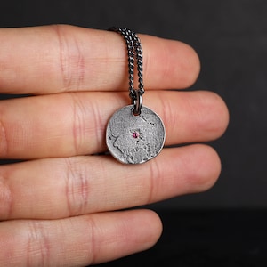 May include: A silver necklace with a round pendant featuring a textured surface and a small pink gemstone in the center.