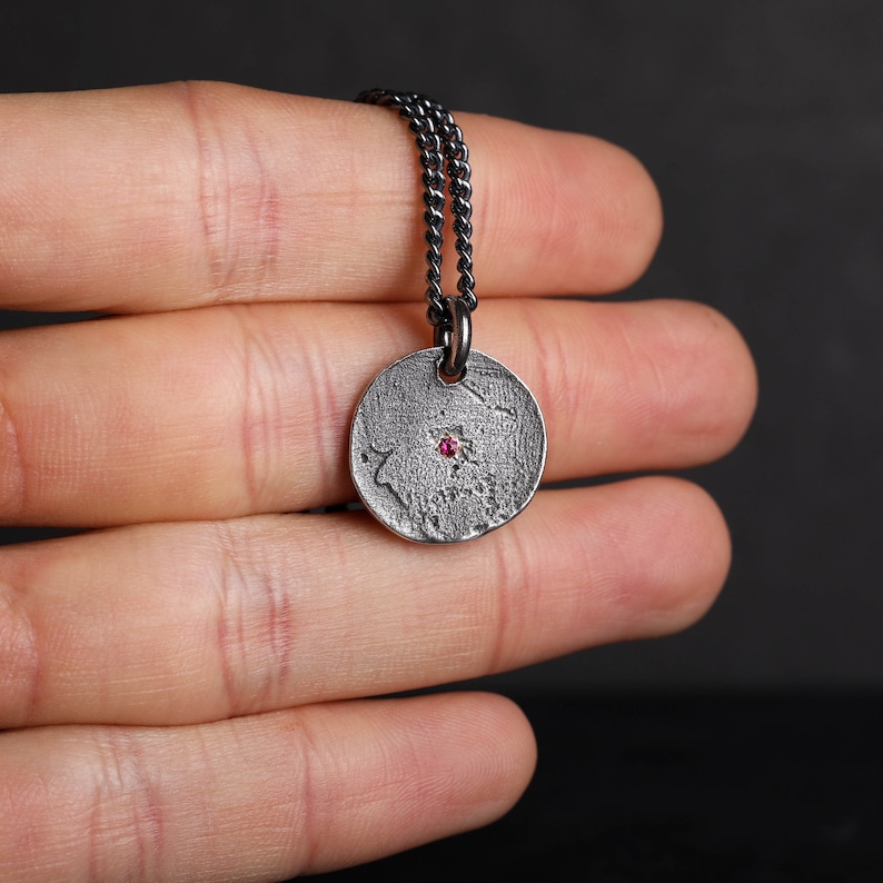 May include: A silver necklace with a round pendant featuring a textured surface and a small pink gemstone in the center.