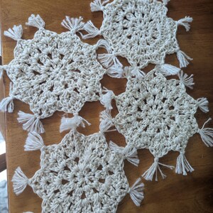May include: Four off-white, crocheted snowflake-shaped coasters with white tassels at each point. The coasters are arranged on a wooden surface. The crochet pattern is visible, and the tassels add a decorative element.