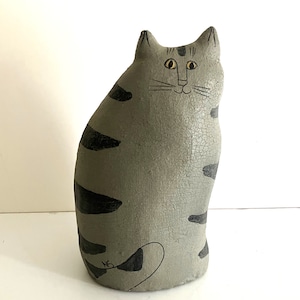 May include: A grey ceramic cat figurine with black stripes. The cat has a simple, cartoon-like face with black eyes and a yellow nose.