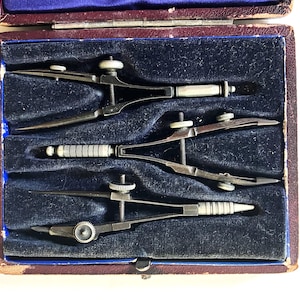 May include: A set of three vintage drafting compasses and dividers, neatly arranged in a velvet-lined case. The tools are made of metal with silver and black accents, showcasing precision engineering. The case is a deep blue and burgundy.