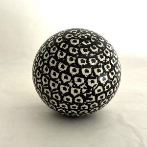 May include: A spherical object featuring a repeating black and white pattern that resembles a dense, textured honeycomb or scale-like design. The surface displays a high-contrast, monochromatic aesthetic with small, irregular shapes covering the sphere.