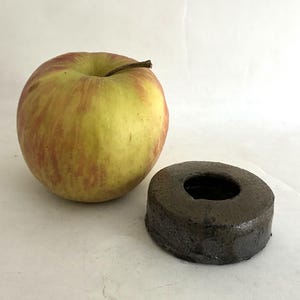 May include: A round, dark brown ceramic object with a central hole, featuring a textured, uneven surface and a glossy finish, positioned next to a large, round, yellow and red apple with a brown stem for a visual size comparison.