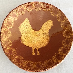 Peut inclure: Une assiette ronde en céramique marron avec une silhouette de poulet dorée au centre. L'assiette a une bordure texturée et tachetée dans des tons de marron et d'or. Idéale pour servir des aliments.