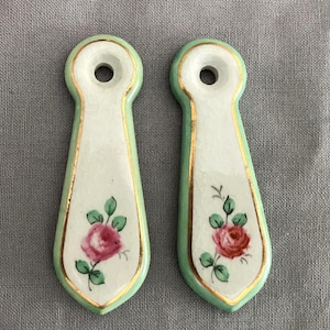 May include: Two white ceramic drawer pulls with green and gold trim. Each pull features a pink rose with green leaves.