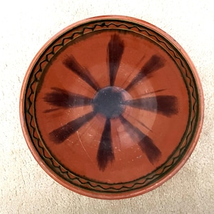 May include: A handmade, terracotta-colored ceramic bowl. The bowl features a dark brown, sunburst-like pattern radiating from a central dark blue circle. The rim is decorated with a black and green wavy design.