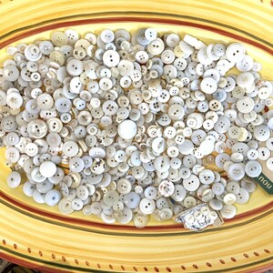 May include: A large collection of white and off-white buttons of various sizes and shapes in a yellow and brown oval dish. The buttons are mostly round, but some are square, oval, and rectangular. There is a small tag with the word "now" printed on it.