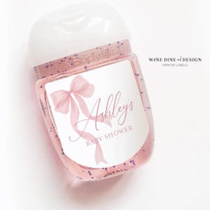 K&ouml;nnte beinhalten: Eine durchsichtige Plastikflasche mit einem wei&szlig;en Pumpspender und einem rosa-wei&szlig;en Etikett. Das Etikett zeigt eine rosa Schleife und den Text "Ashley's Baby Shower".