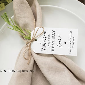 May include: A white tag tied to a beige linen napkin with a sprig of rosemary. The tag reads "Tomorrow is going to be the BEST DAY Ever" with a small black heart. The date "07.16.2021" and the names "CHLOE AND THOMAS" are printed below the text.