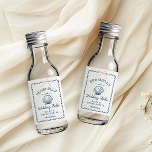 May include: Two clear glass bottles with silver caps. Each bottle has a white label with blue scalloped edges, the words "Seashells and Wedding Bells" and a seashell graphic. The text "Riley's Bachelorette Charleston" is also on the label.