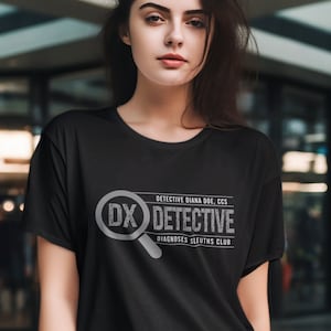 Pode incluir: Camiseta preta com um gráfico cinza com uma lupa e as letras "DX". O texto acima e abaixo diz "DETECTIVE DIANA DOE, CCS" e "DIAGNOSES SLEUTHS CLUB". A camisa é usada com jeans azul claro.